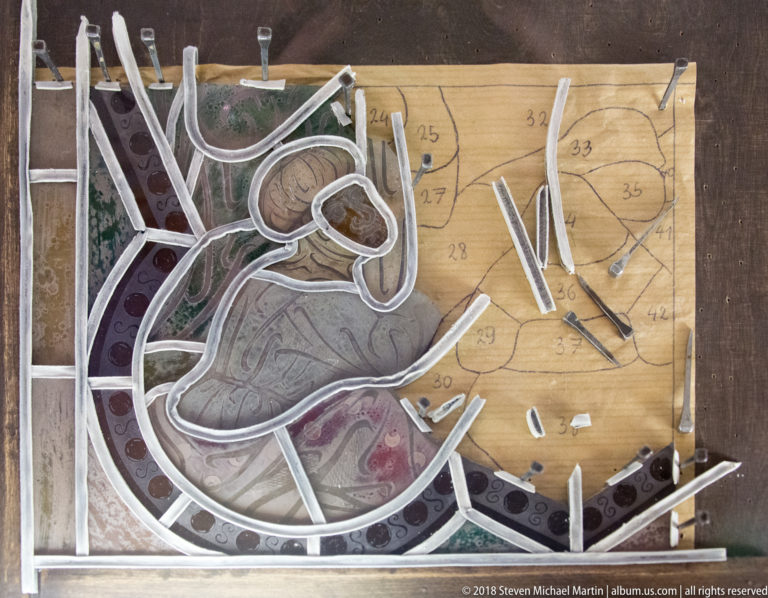 Cut and painted stained-glass pieces aligned over a numbered full-size cartoon during assembly of a figure.