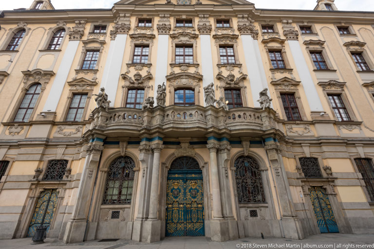 Ornate pale Baroque university façade with central stone balcony, sculptural portal, tall windows, and decorative metal gates.