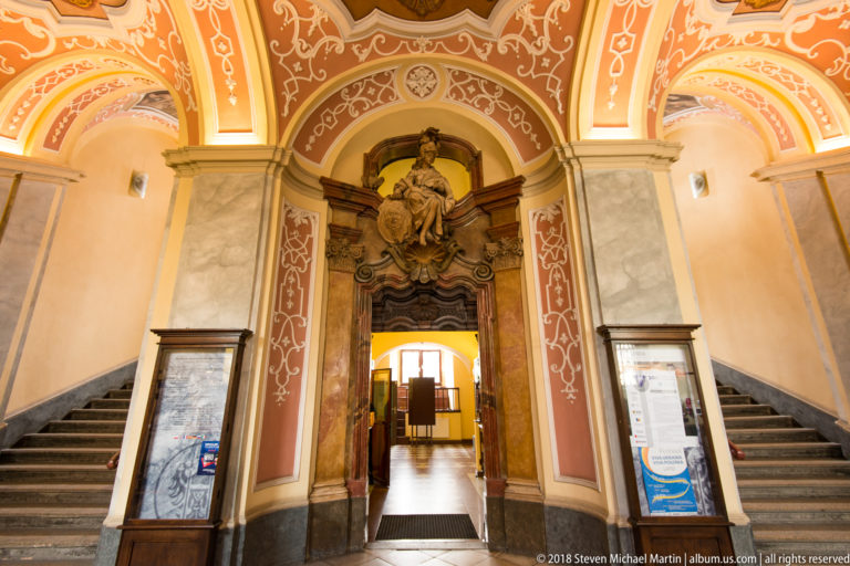 Central Baroque doorway with sculptural overdoor, flanked by two stone stair flights beneath ornate vaulted decoration.
