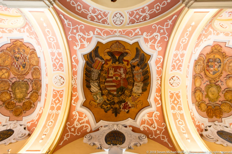 Double-headed eagle with central coat of arms surrounded by heraldic shields and ornate coral-and-white ceiling decoration.