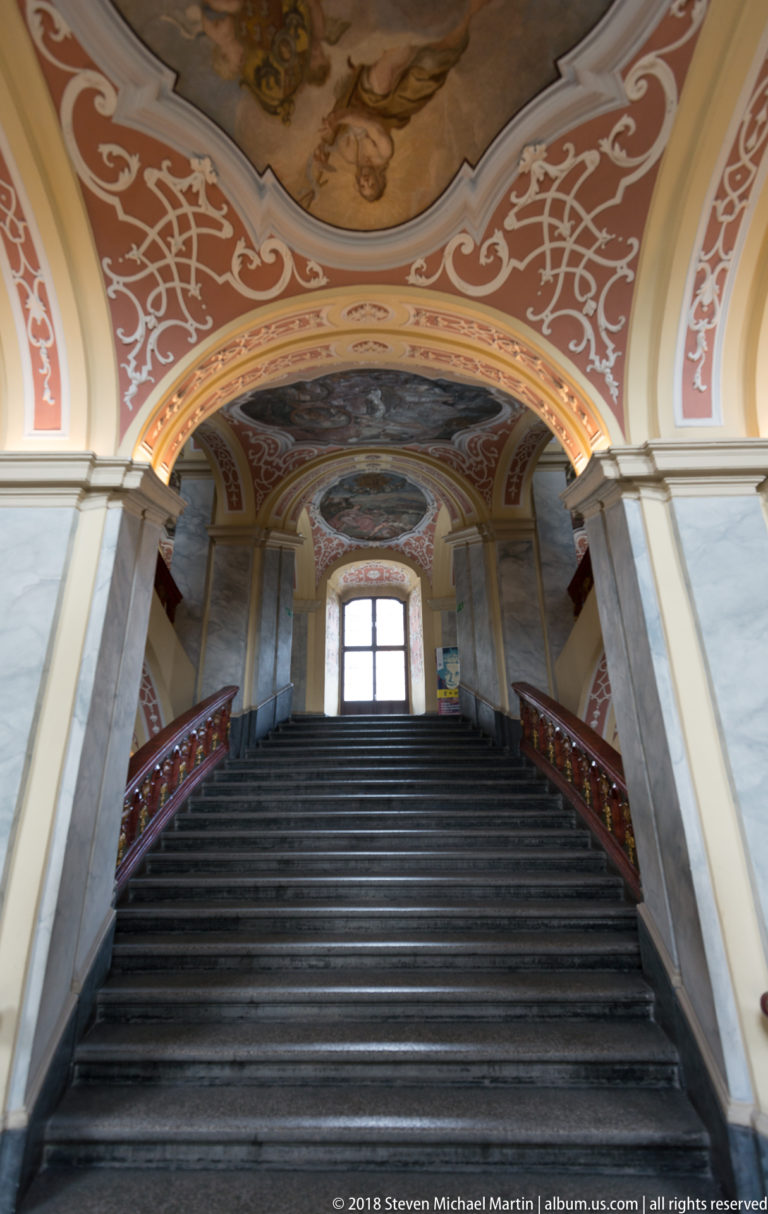 Low centered view up broad dark stone steps between red rails and pale piers beneath decorated vaults.