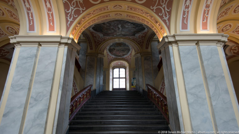 Centered view up a broad dark staircase between red rails and marbleized piers beneath frescoed Baroque vaults.