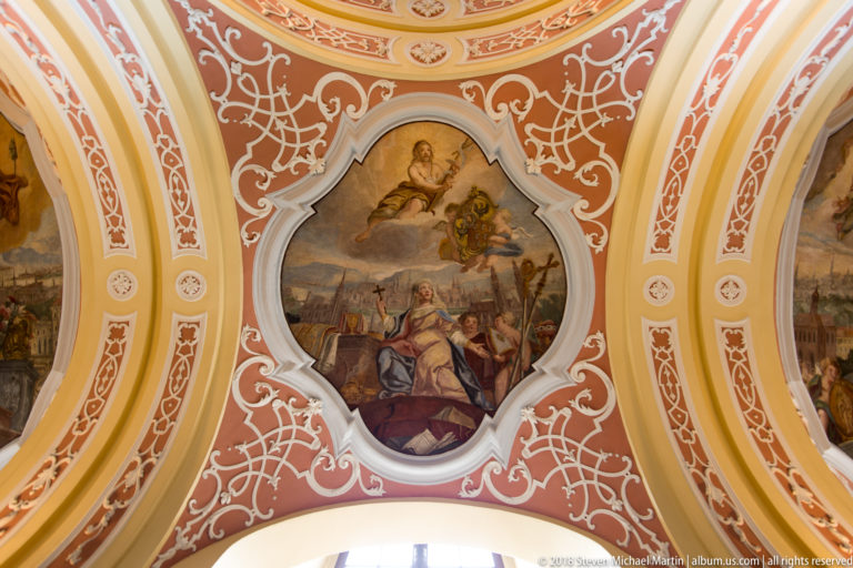 Oval figurative ceiling fresco surrounded by elaborate coral-and-white Baroque scrollwork and adjacent painted panels.