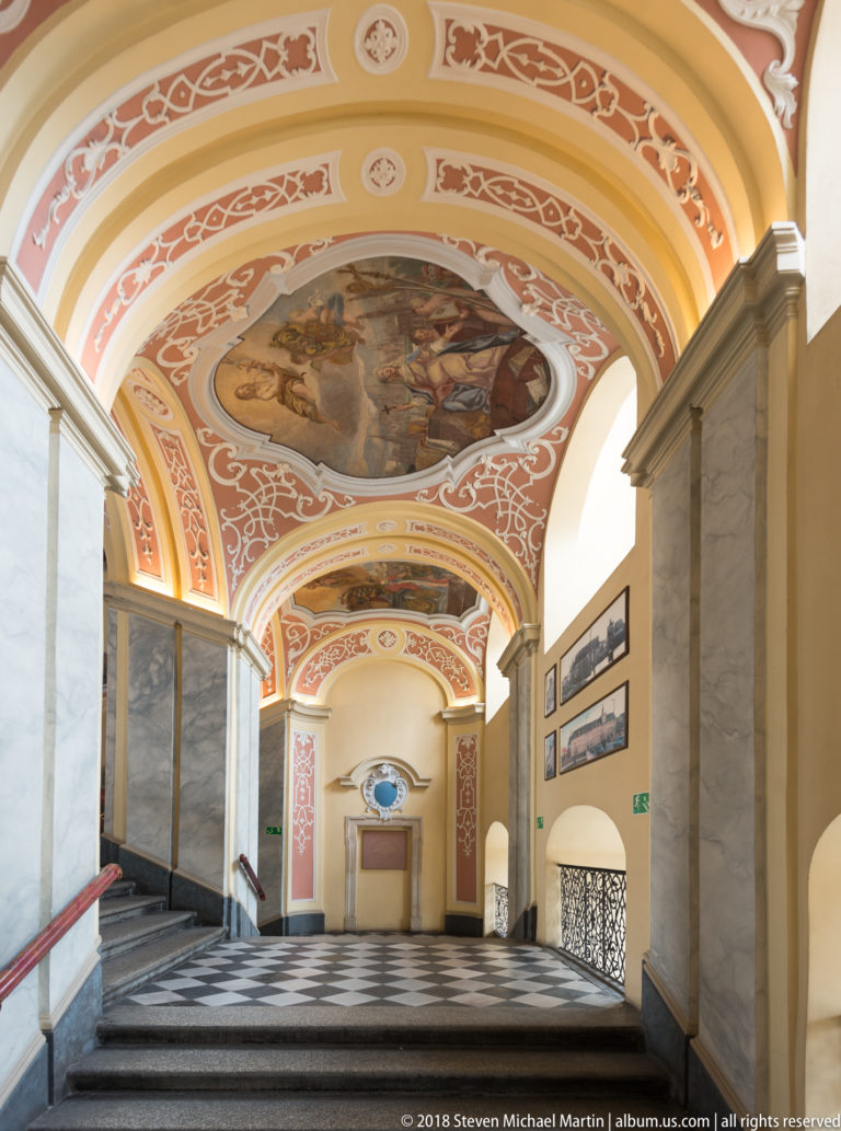Wide stone staircase descending beneath coral-and-white Baroque vaults with ceiling paintings and marbleized wall panels.