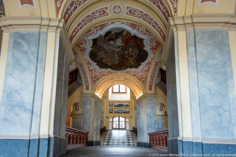 Long Baroque stair hall with painted vaults, marbleized piers, red balustrades, and black-and-white flooring.
