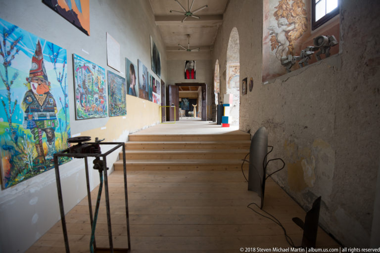Long stepped gallery corridor with colorful contemporary paintings, small sculptures, rough walls, and exposed historic masonry.