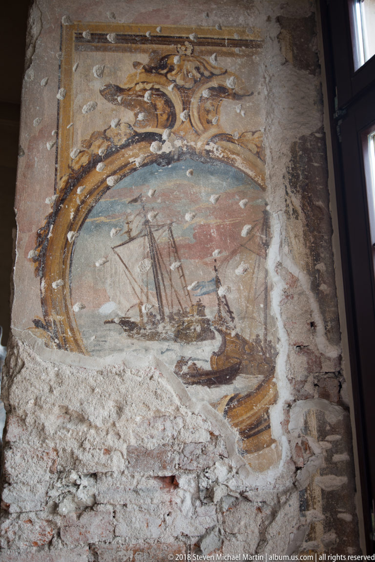Fragmentary painted cartouche with a multi-masted sailing ship exposed amid rough plaster and brickwork.