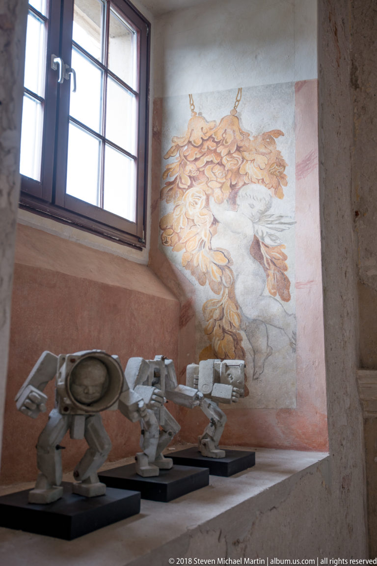 Small pale contemporary sculptures on black plinths below a window and a large ocher wall painting.