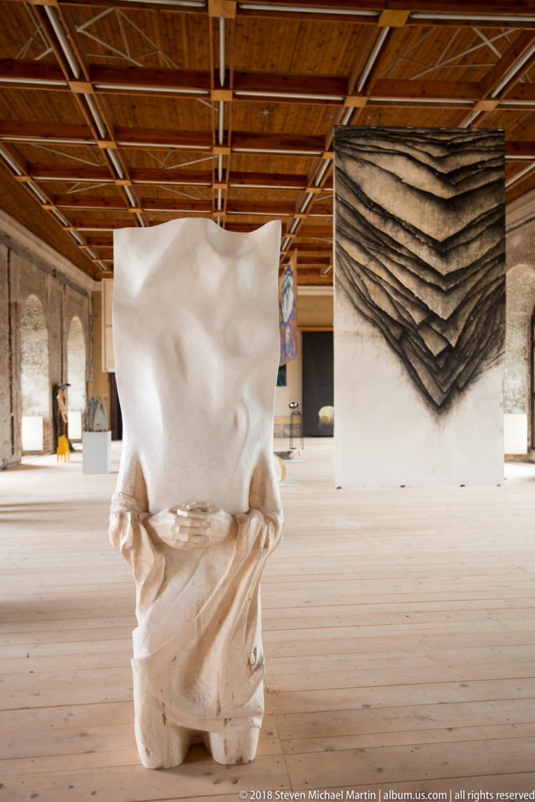 White abstract sculpture with folded lower forms centered in a large gallery beneath a timber ceiling.