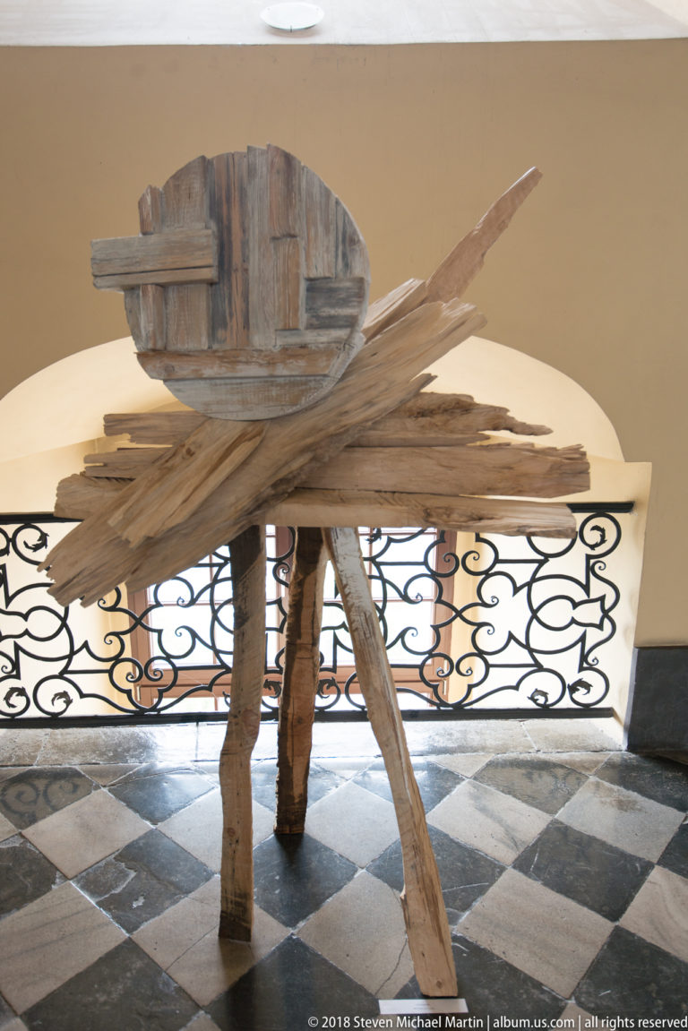 Rough wooden assemblage with projecting boards and a rounded upper form standing on four legs over a checkered floor.