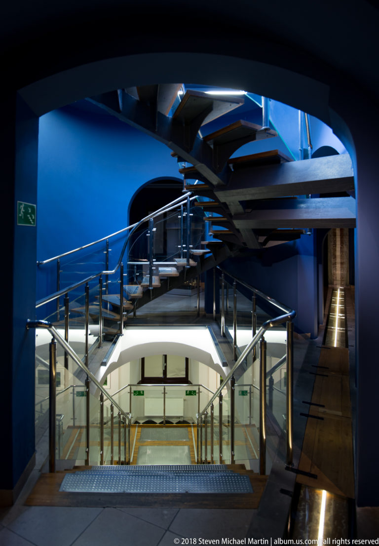 Spiral staircase, blue walls, glass railings, and illuminated walkways framed by a dark curved opening.
