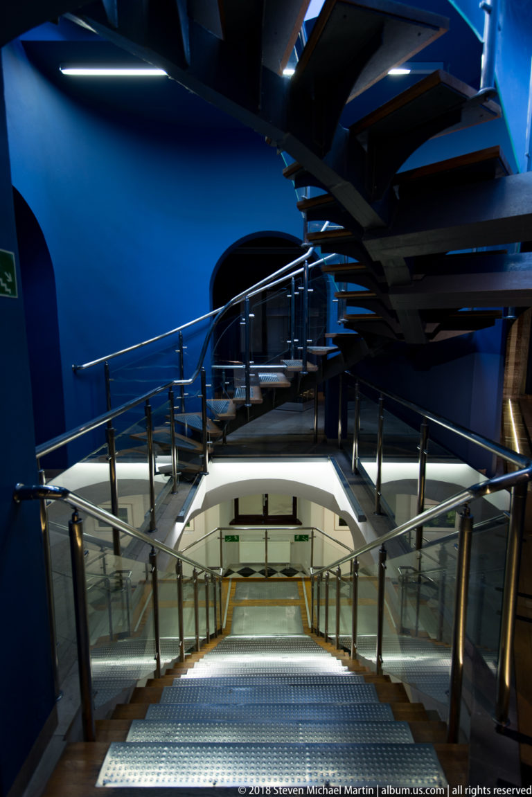 Dark spiral staircase curving above glass railings and a bright lower landing inside a deep-blue interior.