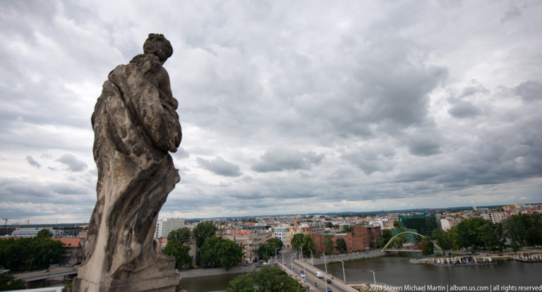 Weathered stone rooftop figure in profile above streets, trees, and distant Wrocław neighborhoods under heavy clouds.