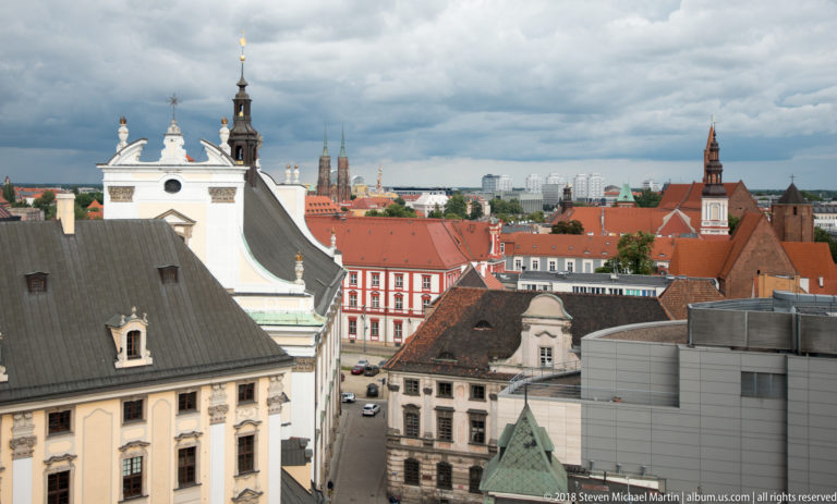Ornate pale buildings and varied roofs in the foreground with Wrocław church towers across the skyline.