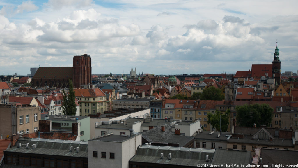 Elevated view over Wrocław buildings toward red roofs and multiple church towers beneath a cloudy sky.
