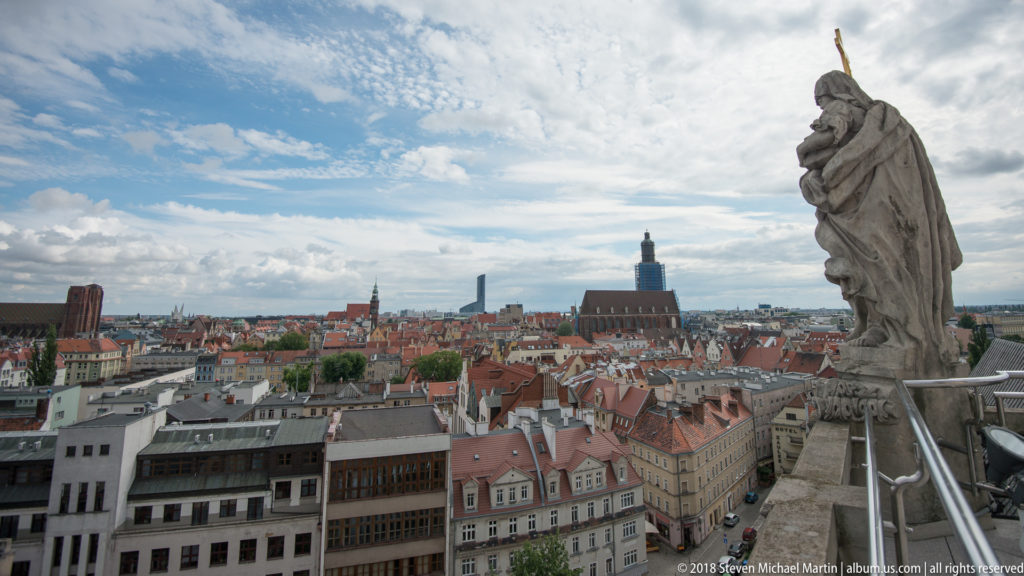 Stone rooftop figure at right overlooking dense historic buildings and church towers across central Wrocław.