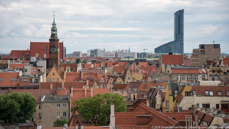 Panorama of Wrocław red-tiled roofs, a historic church tower, and the distant modern Sky Tower.