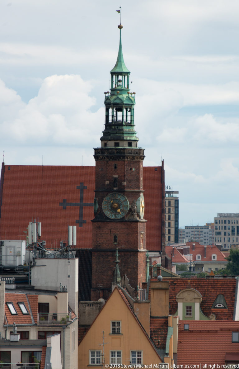 Brick clock tower topped by a green copper belfry and spire rising above central Wrocław rooftops.