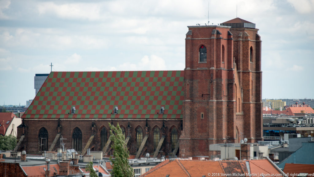 Large Gothic brick church with a tall square tower and patterned red-and-green roof amid Wrocław rooftops.