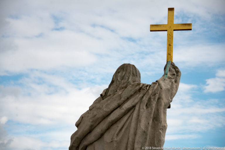 Close upward view of a weathered draped stone rooftop figure holding a gilded cross against the sky.
