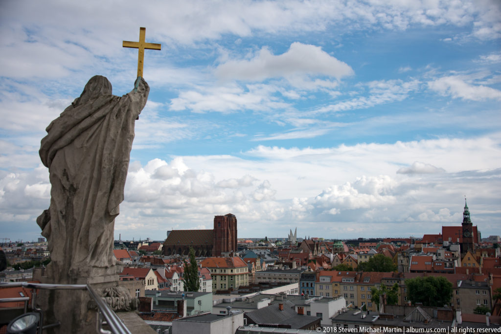 Stone rooftop figure holding a gilded cross above the red roofs and church towers of central Wrocław.