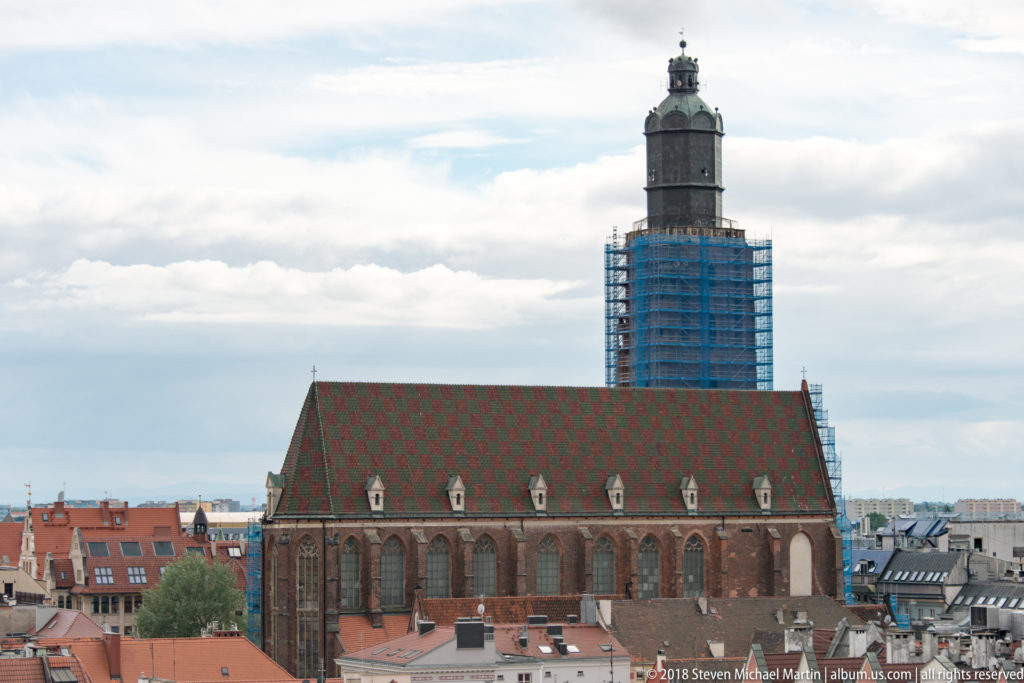 Large brick Gothic hall church with steep patterned roof and tall square tower wrapped in blue scaffolding above Wroclaw rooftops.