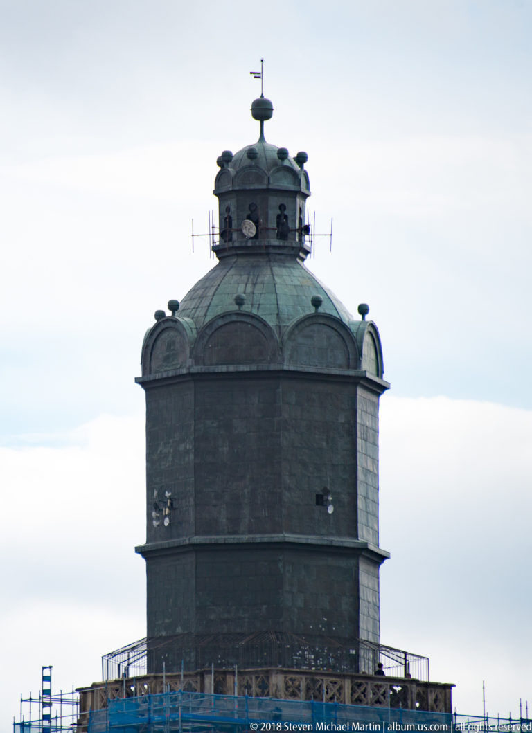 Dark polygonal tower with rounded copper roof, small openings, weather vane, and maintenance platform against a pale sky.