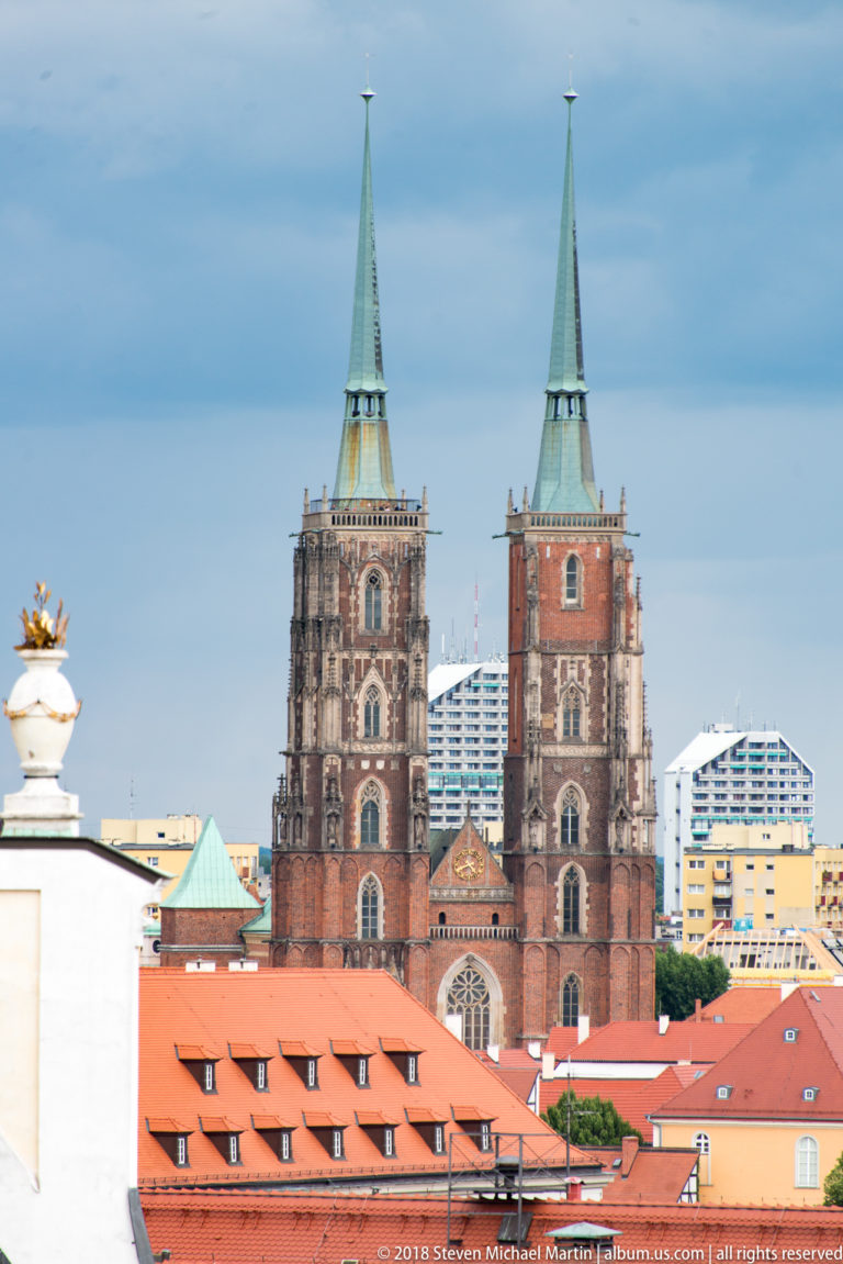 Twin brick-and-stone towers and green spires of Wroclaw Cathedral rising above red-tiled roofs and modern buildings.