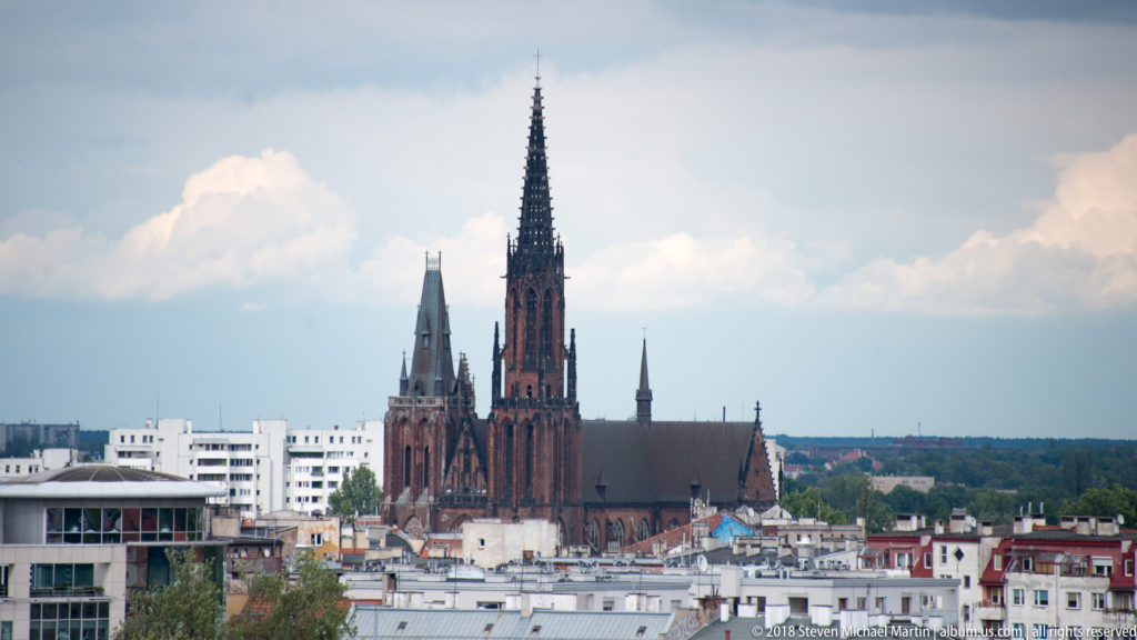 Red-brick Gothic church with a very tall openwork spire rising above Wroclaw rooftops beneath a bright cloudy sky.