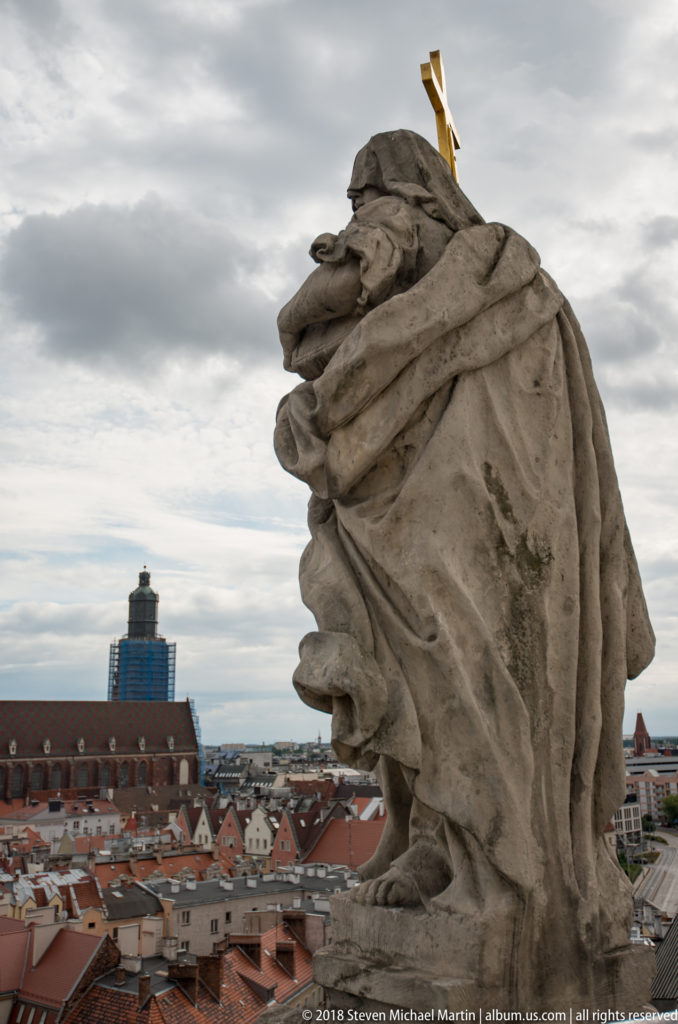 Weathered draped stone figure seen from behind on a rooftop, with a gilded cross-like element and Wroclaw skyline beyond.