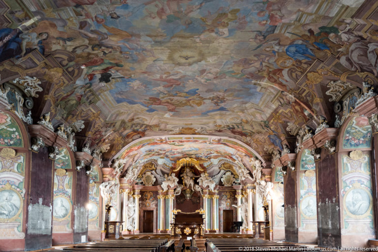 Centered Aula Leopoldina interior with frescoed ceiling, portrait-lined walls, marbled supports, and ornate ceremonial dais at the far end.