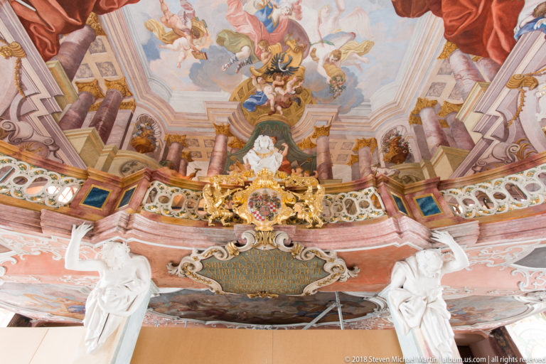Upward view of Aula Leopoldina gallery with Latin inscription cartouche, white statues, gilded heraldry, and ceiling frescoes.