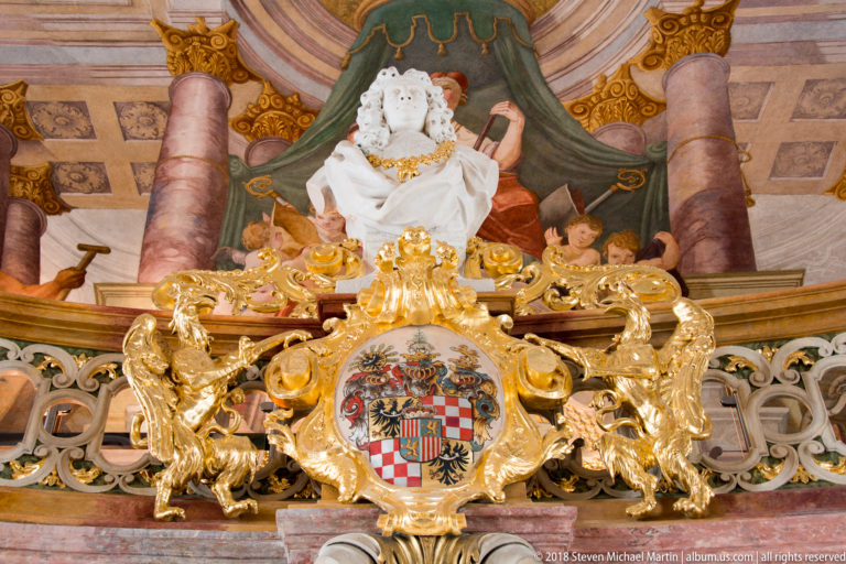 Crowned white bust above a multicolored quartered coat of arms surrounded by gilded scrollwork in Aula Leopoldina.
