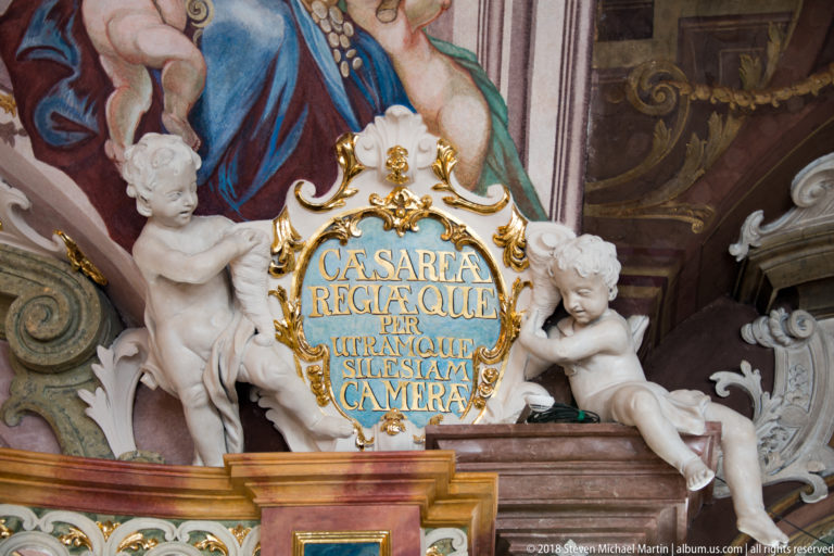 Blue-and-gold Latin inscription cartouche flanked by two white putti amid painted and gilded Baroque ornament.