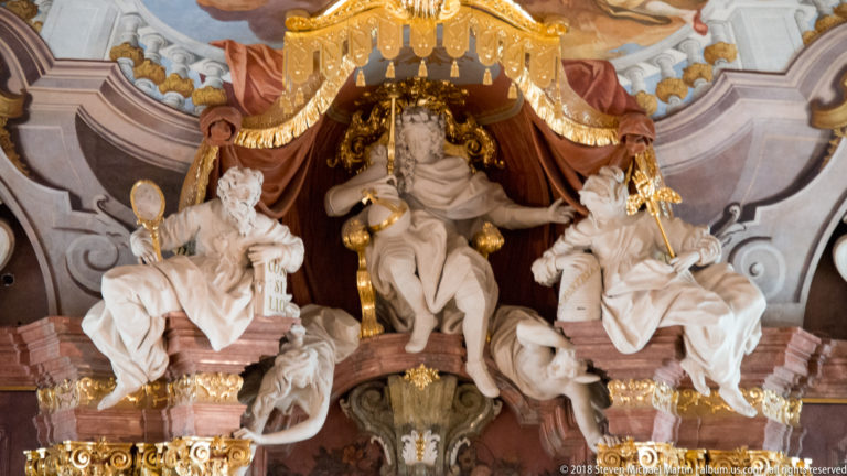 Seated central figure beneath a gilded canopy, flanked by two large white allegorical statues and smaller sculptural figures in Aula Leopoldina.