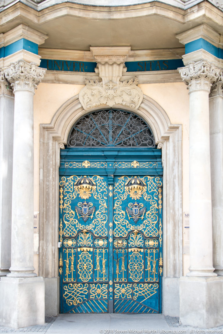 Close frontal view of ornate blue wrought-iron double doors with gilded scrollwork beneath an arched grille.