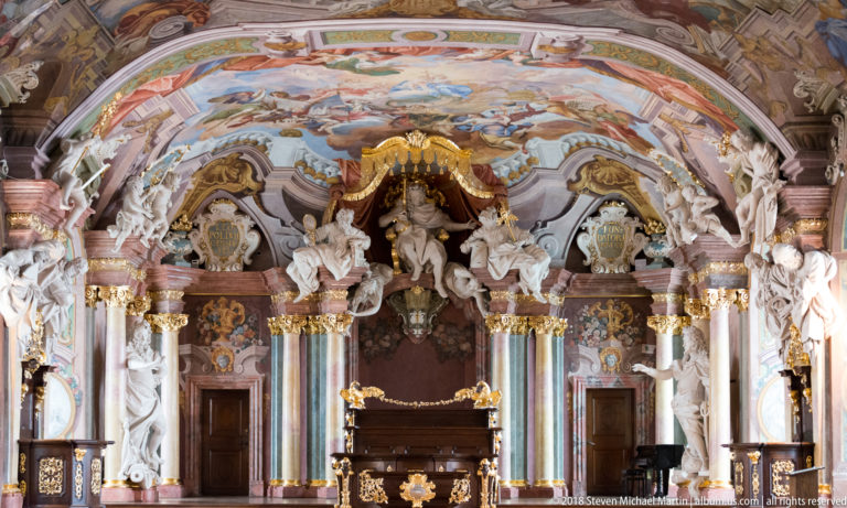 Frontal view of Aula Leopoldina’s ceremonial dais beneath a frescoed vault, marbled columns, white statues, gilding, and central canopy figure.