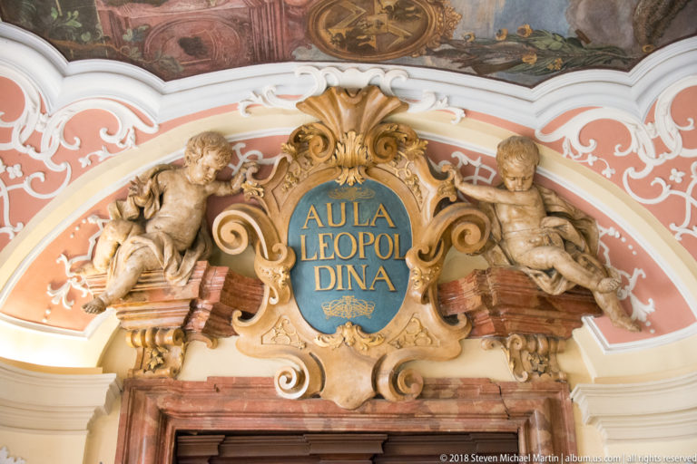 Blue “AULA LEOPOLDINA” cartouche with gilded frame and two reclining sculpted figures above a doorway.