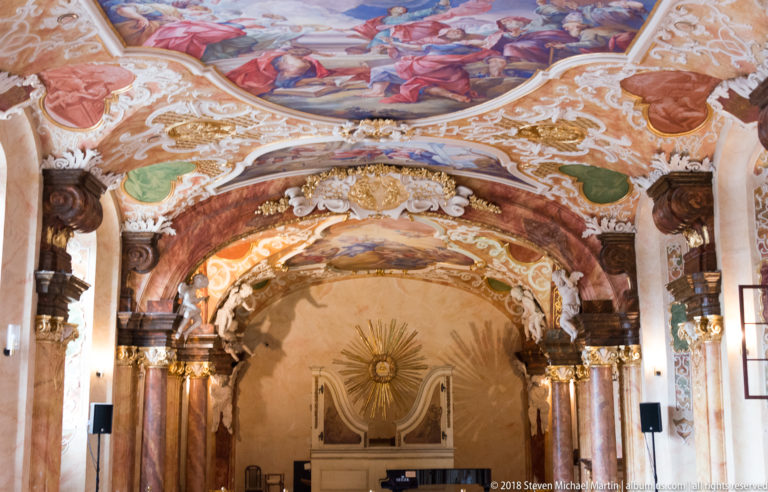 Oratorium Marianum interior with marbled columns, gilded details, painted vaults, white stucco, and a radiant eye motif at the far wall.