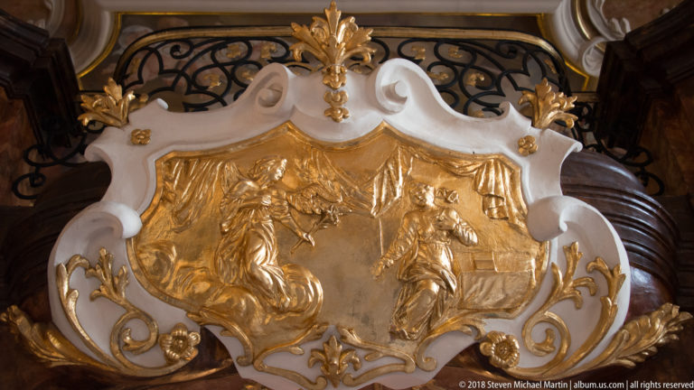White-and-gold scrolling cartouche containing a gilded two-part relief with figures, buildings, foliage, and landscape motifs.