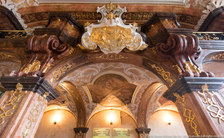 Ornate Baroque interior bay with marbled columns, gilded scrollwork, painted vaults, three arches, and a heraldic crest above.