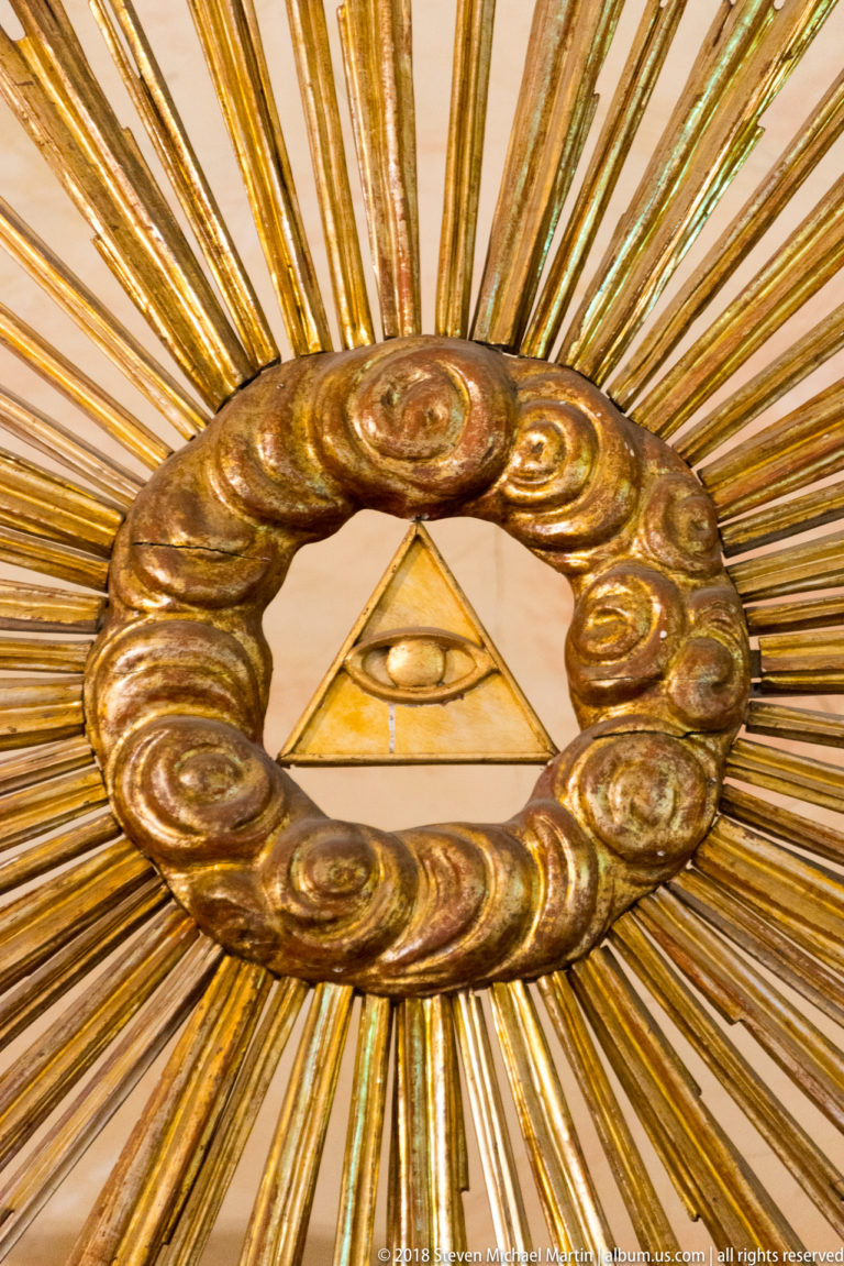 Eye inside a triangle at the center of a gilded sunburst surrounded by a ring of sculpted clouds.