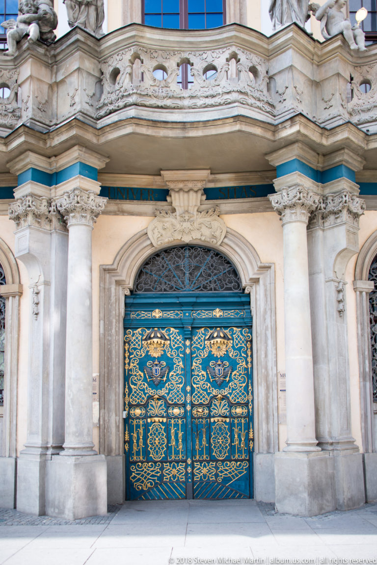 Blue-and-gold wrought-iron double doors framed by pale stone columns and a curved Baroque entrance surround.