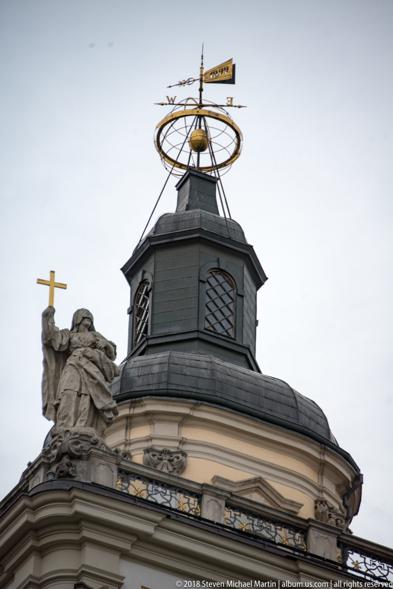 Curved university roof pavilion topped by an armillary-sphere-like metal finial beside a stone figure holding a cross.