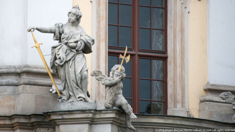 Stone female statue holding a staff beside a small child, mounted on the University of Wroclaw façade near a tall window.