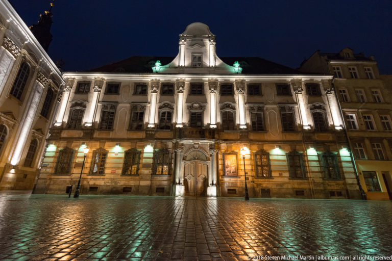 Floodlit University of Wroclaw façade with central entrance, roof pavilion, and reflections on wet cobblestones at night.