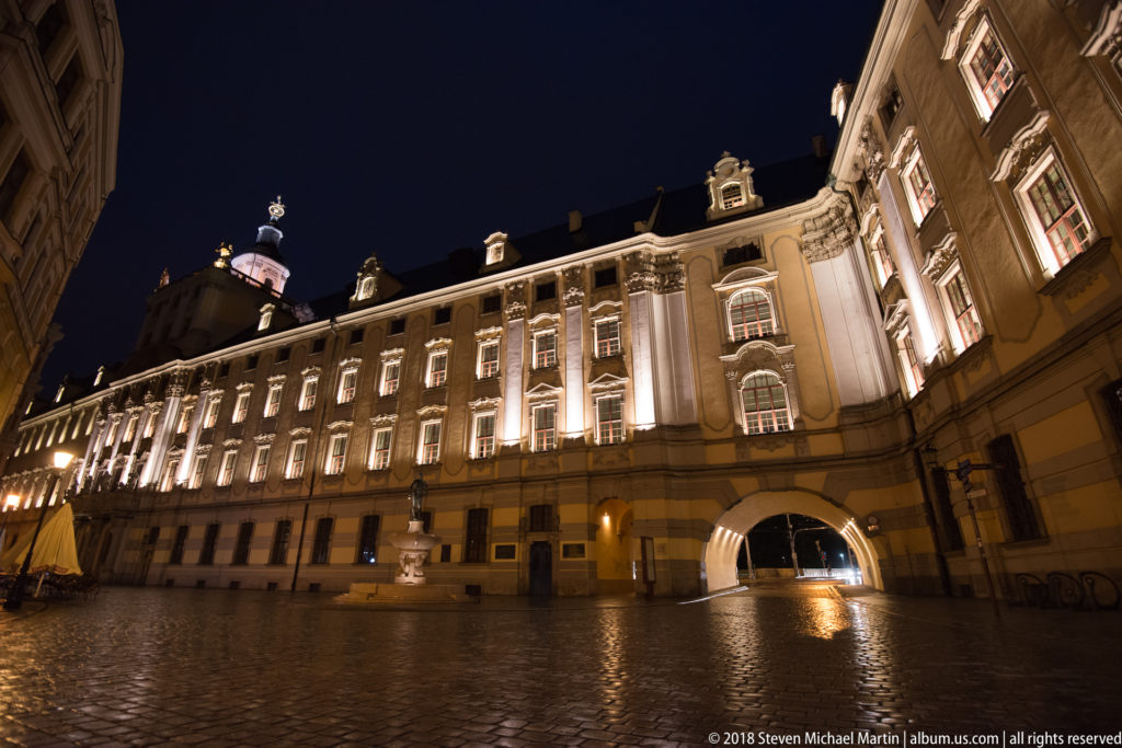 Long illuminated University of Wroclaw façade at night, viewed obliquely across wet cobblestones beneath a dark sky.