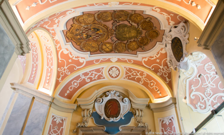 Large heraldic ceiling cluster with many shields and a crowned central coat of arms amid ornate coral-and-white Baroque decoration.