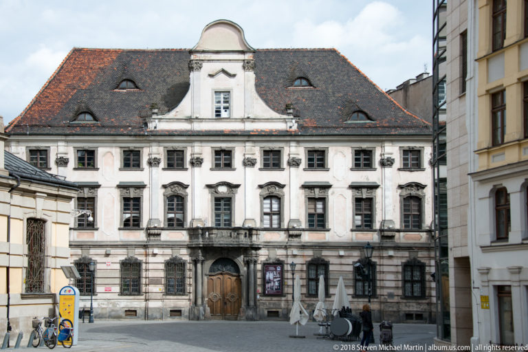 Pale historic building with steep tiled roof, curved central gable, rows of windows, and small balcony facing a paved square.