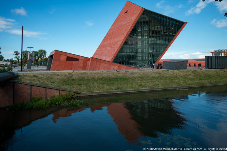 Wide view of the red and glass Museum of the Second World War beside a canal, with its tower reflected in the water.